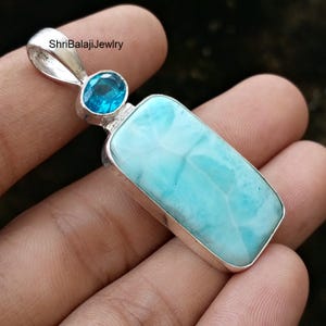 Puede incluir: Un colgante rectangular con una piedra de larimar azul claro y una piedra preciosa azul más pequeña. El colgante tiene un engaste de metal plateado y un lazo para una cadena. El texto "ShriBalajiJewlry" es visible.
