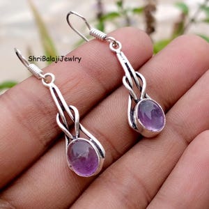 May include: Silver-toned dangle earrings with oval amethyst gemstones. The earrings have a decorative frame and hook closures. The design includes a twisted element. The text "ShriBalajiJewlry" is visible. The earrings are suitable for pierced ears.