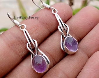 May include: Silver-toned dangle earrings with oval amethyst gemstones. The earrings have a decorative frame and hook closures. The design includes a twisted element. The text "ShriBalajiJewlry" is visible. The earrings are suitable for pierced ears.