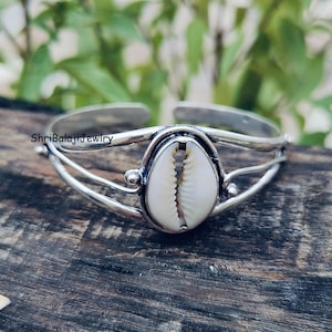 May include: A silver-toned cuff bracelet featuring a white and tan cowrie shell centerpiece. The open-ended bracelet has a simple, elegant design with the text "ShriBalajiJewlry" visible.