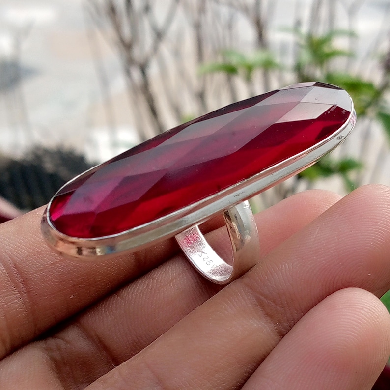 May include: A large, rectangular, faceted, deep red gemstone ring with a silver band. The gemstone is set in a silver bezel and has a long, oval shape. The ring is a statement piece of jewelry.