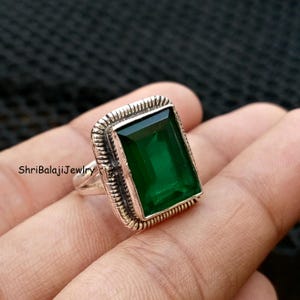 Op de afbeelding: Een zilveren ring met een grote, rechthoekige smaragdgroene edelsteen. De ring heeft een gedetailleerde, getextureerde rand rond de steen. De ring wordt op een hand getoond.