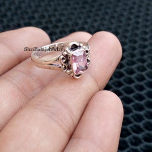 May include: A silver ring with a rectangular, light pink gemstone. The ring has an ornate, filigree design around the gemstone and on the shoulders. The text "ShriBalajiJewlry" is visible on the ring.
