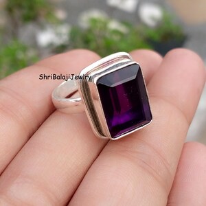 May include: A silver ring featuring a large, rectangular, deep purple gemstone. The gemstone is set in a silver bezel. The ring is displayed against a blurred background. The text "ShriBalajiJewlry" is visible on the ring.