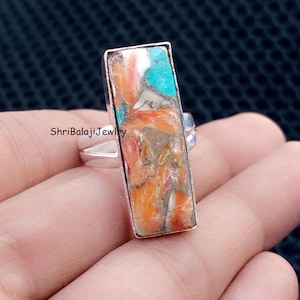 May include: A rectangular silver ring featuring a vibrant, multi-colored stone. The stone displays a mix of orange, turquoise, white, and brown hues. The ring's design is simple, with a polished silver band and a rectangular setting for the stone. The text "ShriBalajiJewlry" is visible.