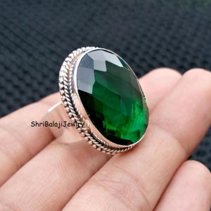 May include: A silver ring featuring a large, faceted, emerald-green gemstone. The oval-shaped stone is the focal point, set in a silver band with a textured, rope-like border. The text "ShriBalajiJewiry" is visible.