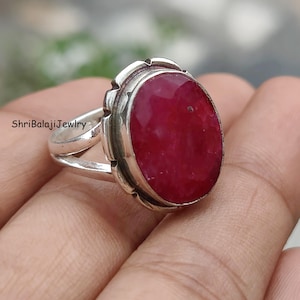 Può includere: Un anello d'argento con una grande gemma di rubino ovale. Il rubino è di un colore rosso intenso ed è incastonato in una lunetta d'argento decorativa. L'anello ha un design a gambo diviso. Il testo "ShriBalajiJewlry" è visibile.