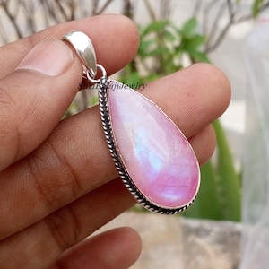 Op de afbeelding: Een druppelvormige hanger met een roze en iriserende edelsteen. De edelsteen is gezet in een zilverkleurig metalen frame met een gedraaid touwontwerp rond de rand. De hanger heeft een zilveren beugel.