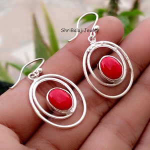 May include: A pair of silver earrings with a red oval stone set in the center of a double oval design. The earrings have a hook closure. The text "ShriBalajiJewlry" is visible on the image.