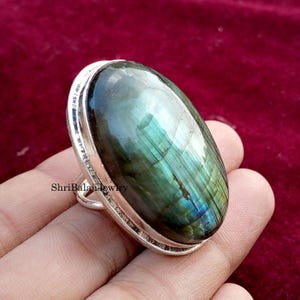May include: A large oval-shaped ring featuring a polished labradorite gemstone. The stone displays iridescent blue, green, and gray hues. The ring has a silver-colored metal band and setting. The jewelry is displayed against a dark red background.