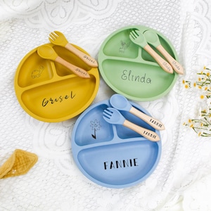 May include: Three divided baby plates in yellow, green, and blue, each with a matching spoon and fork set. The plates are personalized with names in black script. A baby teether is in the lower left corner.