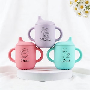 Könnte beinhalten: Drei bunte Baby-Trinkbecher in Rosa, Lavendel und Türkis. Jeder Becher zeigt eine andere Tierillustration und einen Namen: ein Schaf mit "Theo", ein Reh mit "Melina" und eine Ente mit "Joel". Die Becher haben Griffe und einen Ausguss.