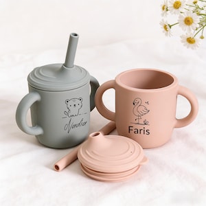 Puede incluir: Dos tazas de bebé de silicona con tapas y asas. Una es gris con un dibujo de oso y el nombre "Nadia". La otra es rosa con un dibujo de flamenco y el nombre "Faris". También se ven una tapa y una pajita rosas.