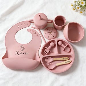 May include: A pink baby feeding set includes a bib, sippy cup, bowl, plate, and utensils. The bib has a duck graphic and the name "Karin". The plate and utensils have wooden handles. The set is made of silicone.