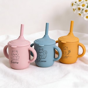 Puede incluir: Tres vasos de silicona coloridos con tapas y asas. Cada taza presenta una ilustración de animal diferente y un nombre: una taza rosa con un zorro y "Siena", una taza azul con una ardilla y "Ronan", y una taza amarilla con un castor y "Eliot".