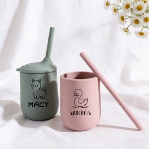 Puede incluir: Dos vasos de silicona con tapas y pajitas. Uno es verde salvia con un gráfico de llama y el nombre "MACY". El otro es rosa con un gráfico de cisne y el nombre "SANTOS". Una pajita rosa descansa contra el vaso rosa. Los vasos están sobre una superficie blanca.