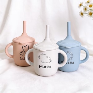 Puede incluir: Tres vasos con boquilla de silicona en colores pastel: rosa, blanco y azul. Cada vaso tiene una tapa con pajita y asa. El vaso rosa tiene un diseño de corazón y las letras "G.I. 3". El vaso blanco tiene un dibujo de animal y el nombre "Maren". El vaso azul tiene un diseño de sol y el nombre "Lena".