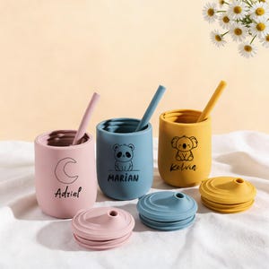 Puede incluir: Tres tazas de silicona de colores con pajitas y tapas. Las tazas son rosa, azul y amarilla, cada una con un gráfico de animal diferente y un nombre. La taza rosa tiene una luna y el nombre "Adriel". La taza azul tiene un panda y el nombre "Marian". La taza amarilla tiene un koala y el nombre "Kelvin".