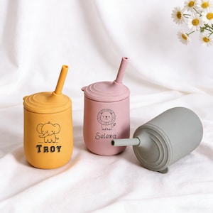 Puede incluir: Tres vasos de silicona coloridos con pajitas. Uno es amarillo con un dibujo de elefante y el nombre "TROY". Otro es rosa con un dibujo de león y el nombre "Selena". El tercer vaso es gris.
