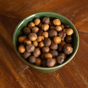 May include: A green ceramic bowl filled with a variety of nuts. The nuts are a mix of brown and tan colors, with some appearing darker than others. The bowl sits on a wooden surface.