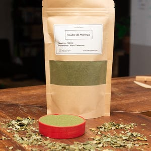 Könnte beinhalten: Eine braune Papiertüte mit Moringa-Pulver, beschriftet mit "Poudre de Moringa". Ein kleiner roter Behälter enthält das grüne Pulver, mit verstreuten Blättern auf einer Holzoberfläche. Die Packung enthält 100 g Moringa.