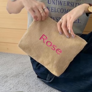May include: A tan corduroy pouch with the name "Rose" embroidered in pink. The pouch is rectangular and appears to be held by a person wearing a gray t-shirt and dark blue jeans.