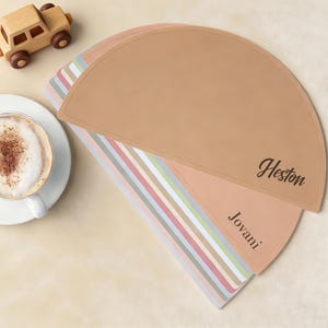 Puede incluir: Un conjunto de manteles individuales semicirculares en tonos beige, rosa y rayas pastel. El mantel superior es beige con el nombre "Heston" impreso. Una taza de café y un coche de juguete de madera también están en la imagen.
