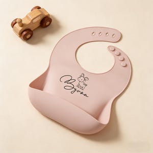 May include: A light pink silicone baby bib with the name "Byron" and a small deer illustration printed in black. The bib has a pocket to catch food and adjustable snaps. A wooden toy car with brown wheels is in the background.