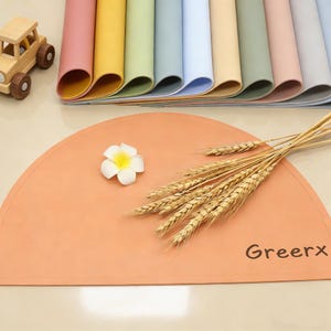 May include: A selection of colourful placemats in shades of pink, yellow, green, and blue. A semi-circular peach-coloured placemat with the word "Greerx" is in the foreground, decorated with a white flower and wheat stalks. A wooden toy car is in the upper left corner.