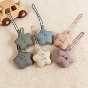 May include: A collection of six star-shaped baby pacifier holders in various pastel colors, each with a loop and a different engraved design. The holders feature a bottle, a lion, a bear, a dinosaur, and the names "Bringy", "Goran", "Alcott", "Hola", and "G.B". A wooden toy car is in the background.