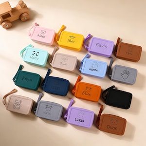 Puede incluir: Una colección de estuches rectangulares y coloridos con correas para la muñeca. Cada estuche es de un color diferente y presenta un nombre o un gráfico, como un oso, un panda o una huella de mano. Un tren de juguete de madera está en la esquina superior izquierda.