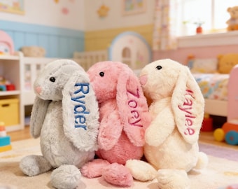May include: Three plush bunny rabbits in gray, pink, and cream colors. Each bunny has a name embroidered on its ear: Ryder, Zoey, and Kaylee. The bunnies are sitting on a light-colored rug in a child's room.
