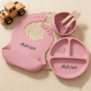 May include: A pink baby feeding set with the name "Adrian" printed on the bib and plate. The set includes a bib, a divided plate with an elephant graphic, a bowl, and a spoon and fork. A wooden toy truck is also in the image.
