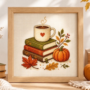 May include: A framed cross-stitch embroidery featuring a stack of three books in green, red, and brown, topped with a cream-coloured mug with a red heart. An orange pumpkin and autumn leaves in red and yellow are arranged around the books.