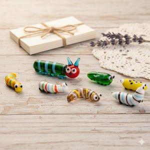 May include: A collection of colorful glass caterpillar figurines. The caterpillars are in various colors, including green, blue, yellow, and white, with striped and dotted patterns. A small stack of cards tied with twine is in the background.