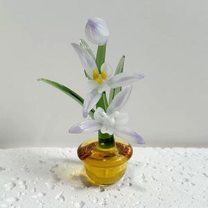 Könnte beinhalten: Ein handgefertigtes Glasblumenarrangement mit weißen und lavendelfarbenen Blütenblättern, gelben Zentren, grünen Stielen und Blättern. Die Blumen stehen in einem kleinen, bernsteinfarbenen Glastopf. Eine zarte und künstlerische Blumenskulptur.