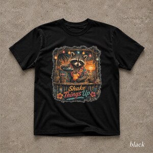 Könnte beinhalten: Ein schwarzes T-Shirt mit einer Grafik eines Waschbären, der ein Blumenhemd und eine Blumenkette trägt und an einer Strandbar einen tropischen Drink mixt. Das Motiv zeigt Lichterketten, einen Sonnenuntergang und den Text "Shake Things Up".