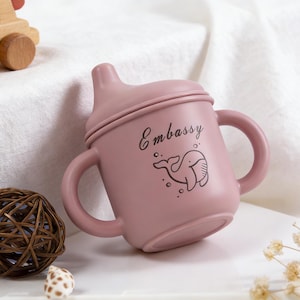 Puede incluir: Un vaso para beb&eacute; de color rosa polvoriento con dos asas y una tapa. La taza tiene la palabra "Embassy" y una ilustraci&oacute;n de ballena. Un coche de juguete de madera, una bola decorativa y flores secas est&aacute;n en el fondo.