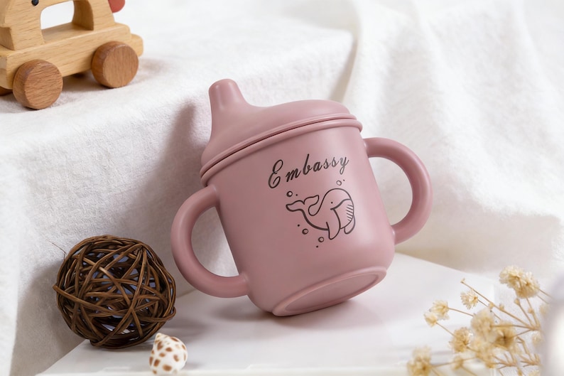 Puede incluir: Un vaso para beb&eacute; de color rosa polvoriento con dos asas y una tapa. La taza tiene la palabra "Embassy" y una ilustraci&oacute;n de ballena. Un coche de juguete de madera, una bola decorativa y flores secas est&aacute;n en el fondo.