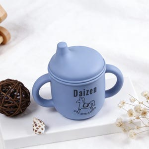 Puede incluir: Una taza para beb&eacute; azul claro con asas y tapa. La taza tiene la palabra "Daizen" y un gr&aacute;fico de caballito balanc&iacute;n. Un caballito balanc&iacute;n de madera y una bola decorativa est&aacute;n en el fondo.