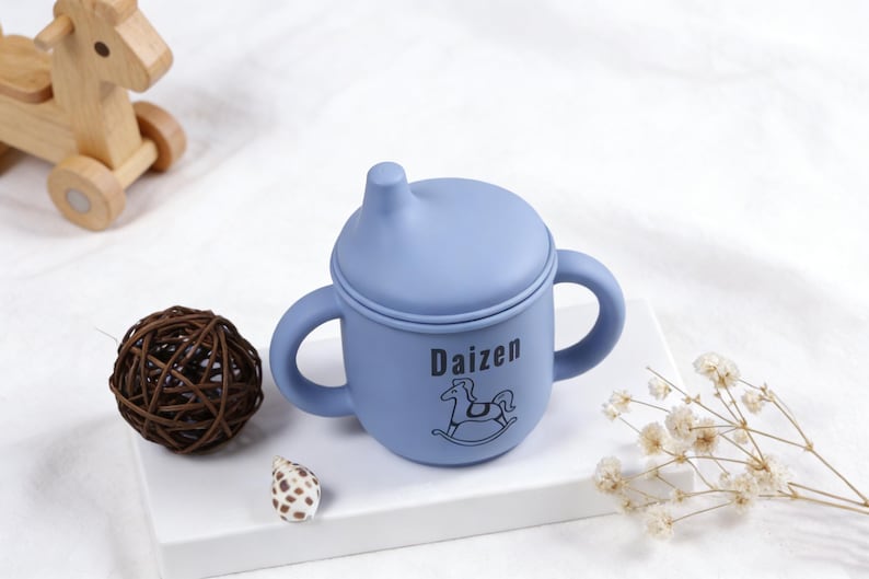 Puede incluir: Una taza para beb&eacute; azul claro con asas y tapa. La taza tiene la palabra "Daizen" y un gr&aacute;fico de caballito balanc&iacute;n. Un caballito balanc&iacute;n de madera y una bola decorativa est&aacute;n en el fondo.