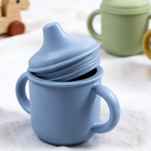 Puede incluir: Una taza para sorber de silicona azul con una tapa a juego, con dos asas para un agarre f&aacute;cil. Otras tazas verdes y amarillas est&aacute;n en el fondo. Tambi&eacute;n se ve un juguete de madera. Este producto para beb&eacute;s est&aacute; dise&ntilde;ado para una alimentaci&oacute;n segura y c&oacute;moda.