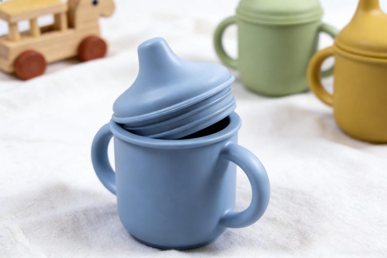 Puede incluir: Una taza para sorber de silicona azul con una tapa a juego, con dos asas para un agarre f&aacute;cil. Otras tazas verdes y amarillas est&aacute;n en el fondo. Tambi&eacute;n se ve un juguete de madera. Este producto para beb&eacute;s est&aacute; dise&ntilde;ado para una alimentaci&oacute;n segura y c&oacute;moda.