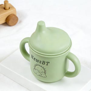 Puede incluir: Un vaso para beb&eacute; de color verde claro con dos asas y una tapa. La taza tiene el texto "NewsBT" y un dise&ntilde;o de sol y nube de dibujos animados. Un tren de juguete de madera est&aacute; en el fondo.