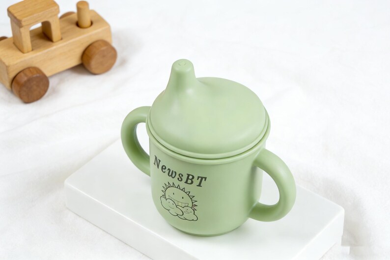 Puede incluir: Un vaso para beb&eacute; de color verde claro con dos asas y una tapa. La taza tiene el texto "NewsBT" y un dise&ntilde;o de sol y nube de dibujos animados. Un tren de juguete de madera est&aacute; en el fondo.
