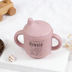 Puede incluir: Un vaso para sorber de color rosa claro con tapa y asas, personalizado con el nombre "Oswald" y una ilustraci&oacute;n de un cohete. El vaso est&aacute; dise&ntilde;ado para beb&eacute;s y ni&ntilde;os peque&ntilde;os. Un juguete de madera y objetos decorativos est&aacute;n en el fondo.