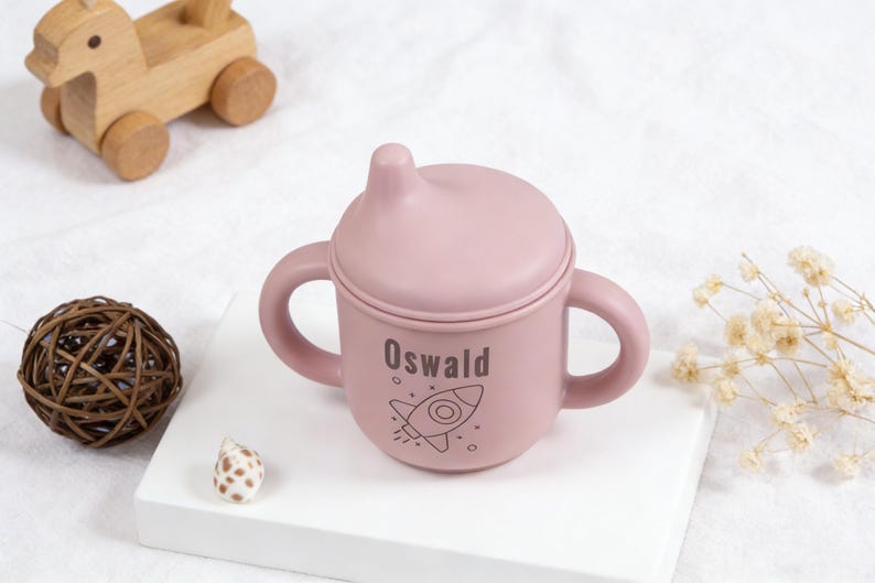 Puede incluir: Un vaso para sorber de color rosa claro con tapa y asas, personalizado con el nombre "Oswald" y una ilustraci&oacute;n de un cohete. El vaso est&aacute; dise&ntilde;ado para beb&eacute;s y ni&ntilde;os peque&ntilde;os. Un juguete de madera y objetos decorativos est&aacute;n en el fondo.