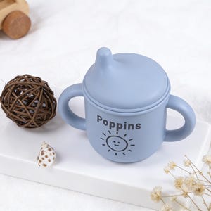 Puede incluir: Una taza para beb&eacute; azul claro con asas y tapa, con la palabra "Poppins" y un sol sonriente. Tambi&eacute;n hay un coche de juguete de madera, una bola decorativa y una concha.