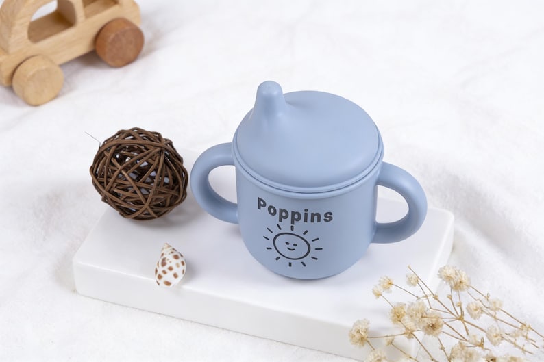 Puede incluir: Una taza para beb&eacute; azul claro con asas y tapa, con la palabra "Poppins" y un sol sonriente. Tambi&eacute;n hay un coche de juguete de madera, una bola decorativa y una concha.