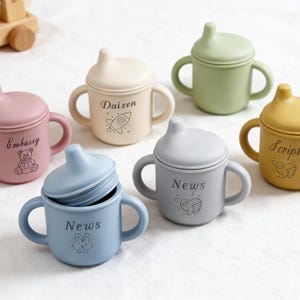 Puede incluir: Colecci&oacute;n de tazas de beb&eacute; de silicona coloridas con asas y tapas. Las tazas son en tonos rosa, crema, verde, amarillo y azul. Cada taza tiene una palabra y un gr&aacute;fico diferentes impresos, como "Embassy" y un osito de peluche.